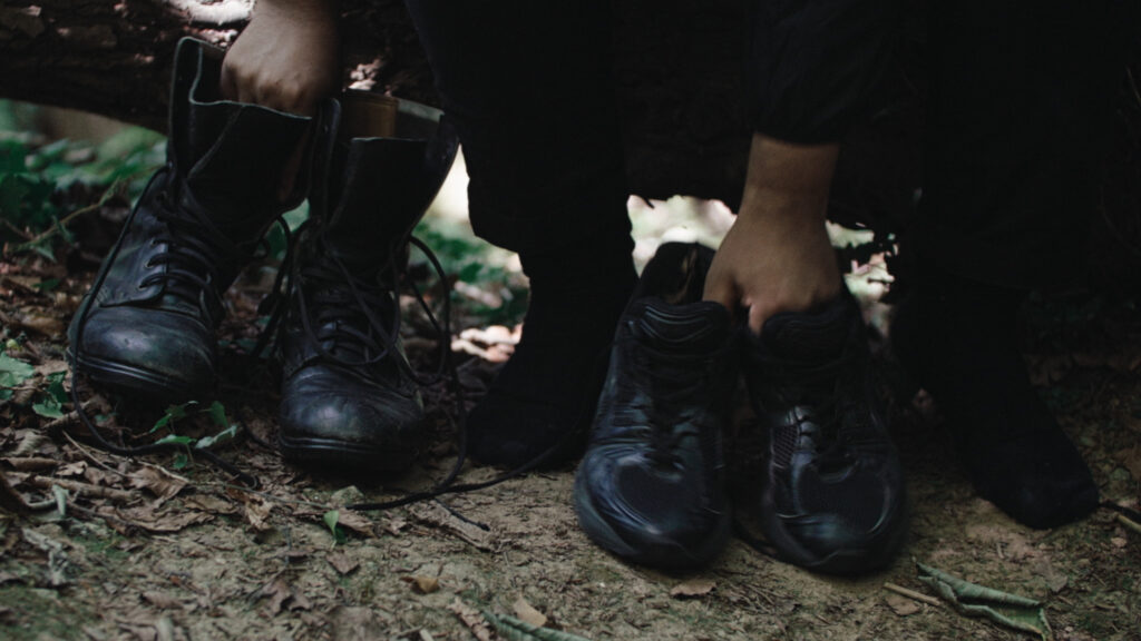 Close up of the feet of a person sitting on a log in the forest with a pair of old fashioned black boots in one hand and a pair of modern black trainers in the other.